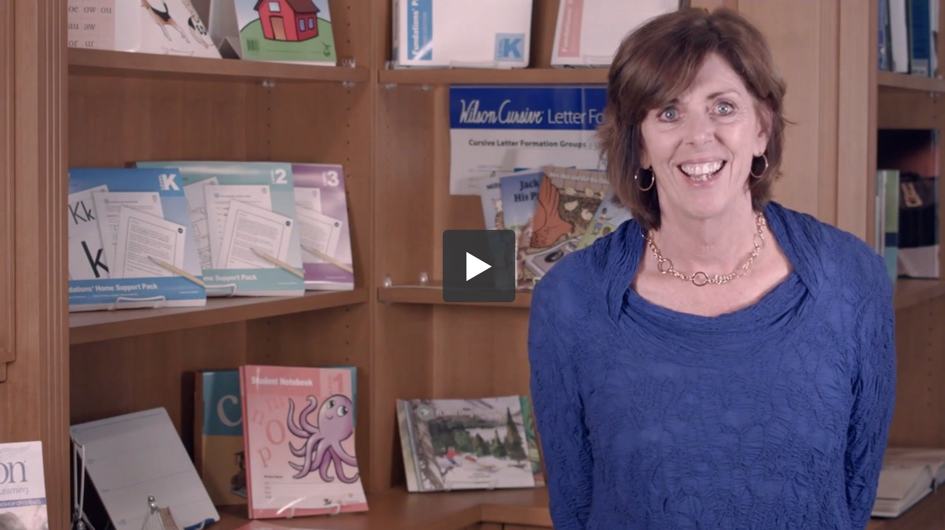 A woman with short brown hair wearing a blue blouse stands smiling in a room with shelves filled with books and educational materials. A video play button is visible at the center of the image, suggesting the frame is from a video.